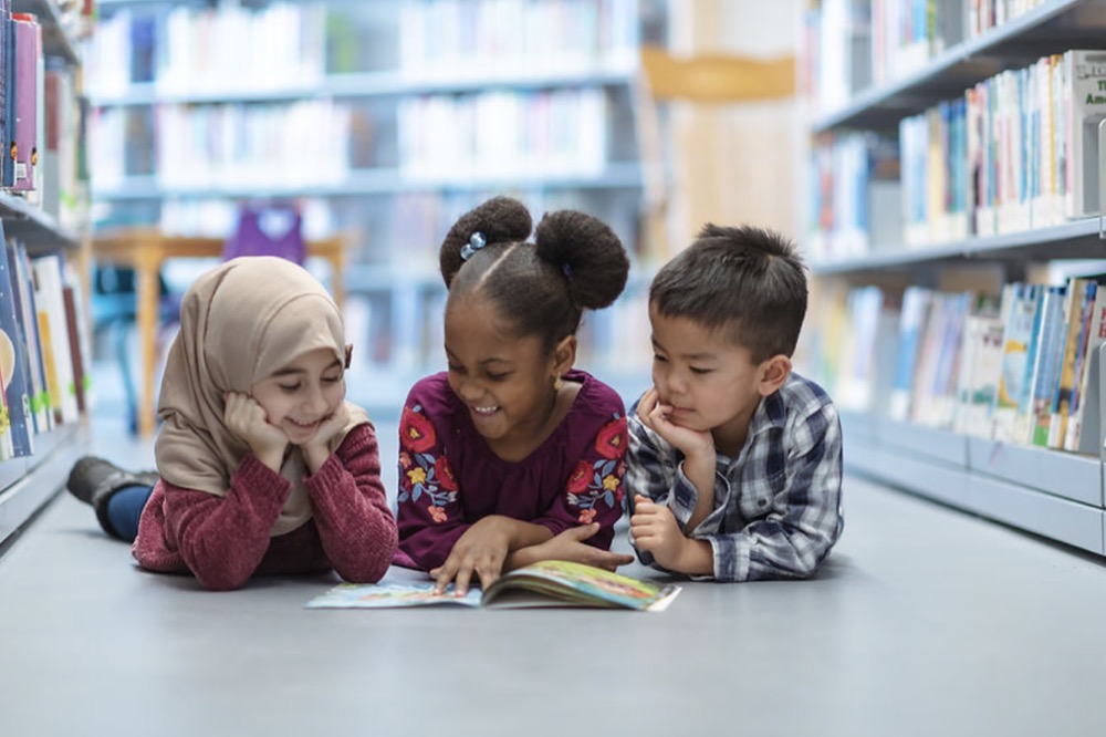 Three racially diverse children lying on the floor of a library looking at a book surrounded by bookshelves.