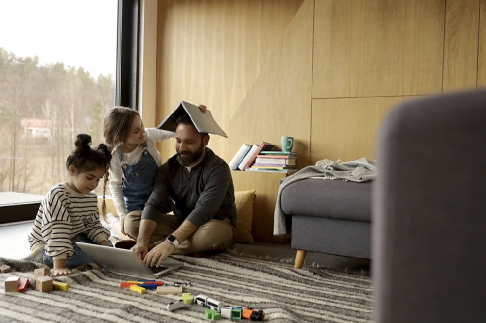 A man typing on a laptop, with two young girls playing with him by putting a book on his head.