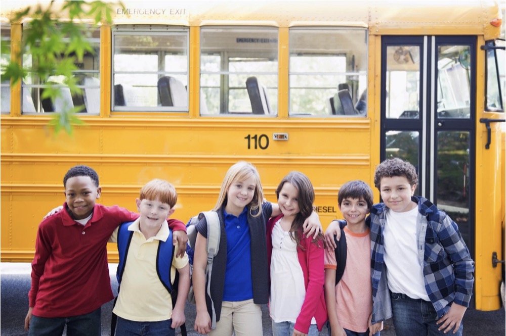 Group of diverse children standing arm-in-arm in front of a yellow school bus.